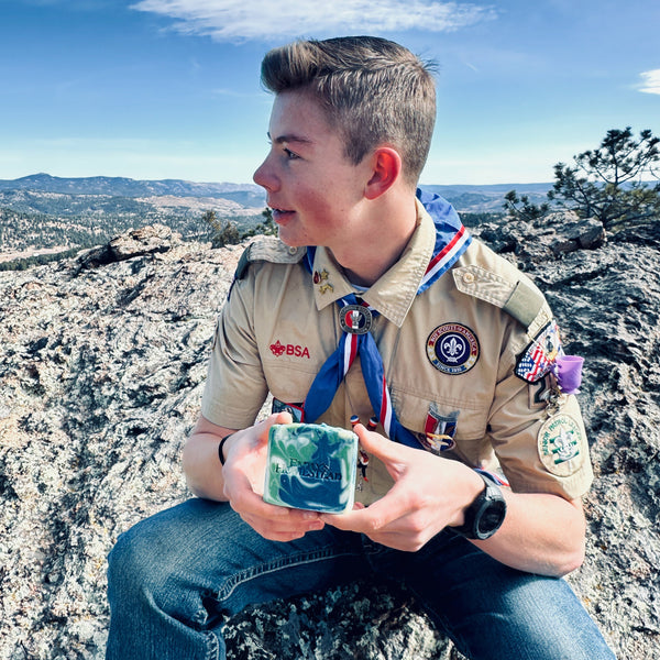 Scout sitting on a rocky outcrop with a scenic background and holding the new scout soap bar